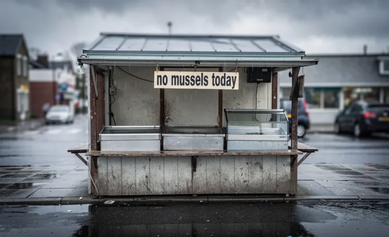 Un stand de marché aux fruits de mer vide avec une pancarte annonçant l'absence de moules, sur un trottoir mouillé sous un ciel printanier nuageux.
