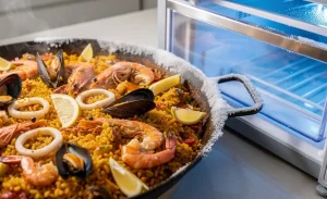 Une paella de fruits de mer colorée repose dans un plat traditionnel à côté d’un tiroir transparent de congélateur, avec de la glace fine visible sur le riz sous une lumière bleutée.