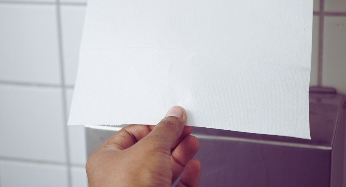 Main tirant une feuille d'essuie-mains en papier depuis un distributeur mural dans des sanitaires carreles